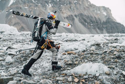 Andrzej Bargiel walking on rocky terrain with skis on shoulder, wearing climbing gear and a helmet in a mountainous landscape.