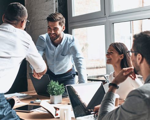 Two men shaking hands at a wooden table, with a woman smiling and another man observing. Laptops and a plant are visible.