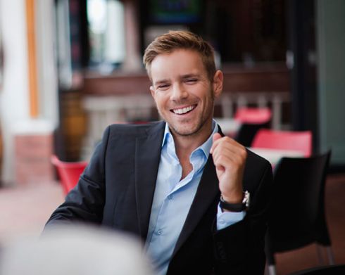 Smiling man in a black suit sitting at a table in a café, with a casual hand gesture and blurred background.