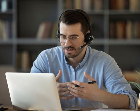 Man wearing a headset gestures while speaking in front of a laptop on a desk, with bookshelves in the background.